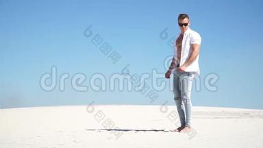 穿牛仔裤的时髦男人，戴着眼镜，穿着白衬衫，光着脚站在沙漠里的沙滩上